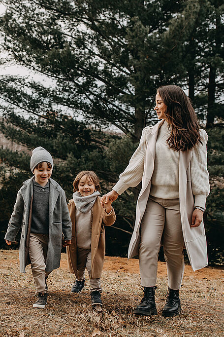 Mother and two children, all wearing coats and scarves, walking hand in hand through a forested area on a cloudy day