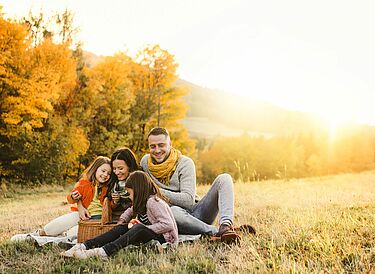 Family of four enjoying a picnic on a sunny autumn day, sitting on a blanket in a field with colorful fall trees in the background