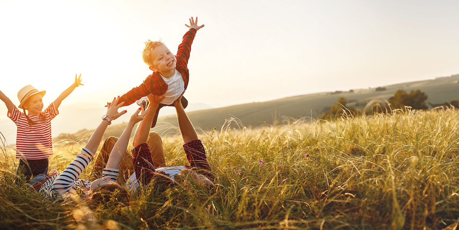 Family playing in a grassy field at sunset, with one adult lifting a joyful child into the air while another child raises their arms in excitement.