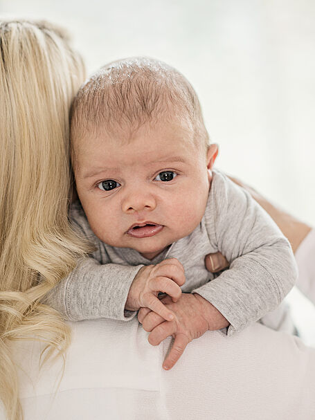 Baby resting on a woman's shoulder, looking directly at the camera, with a calm expression and a light background.