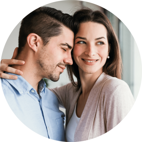 Smiling couple embracing, with the woman looking forward and the man resting his head on her shoulder.